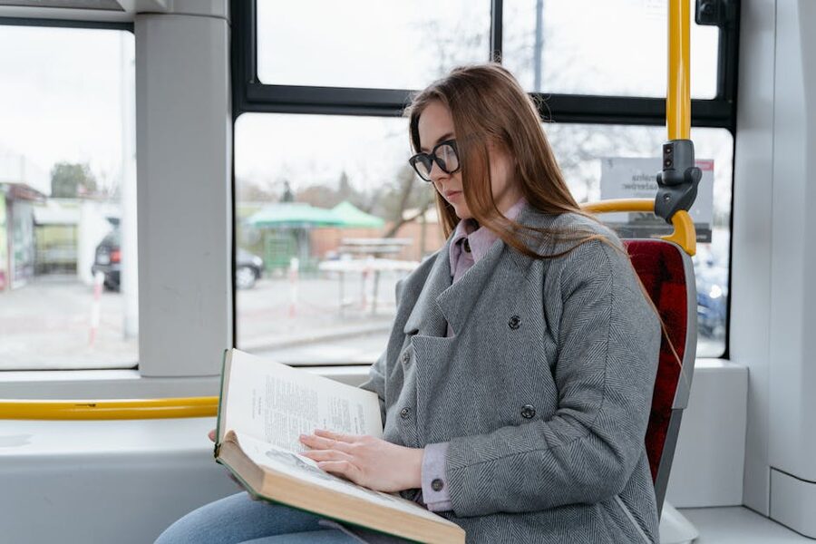 A young woman wearing eyeglasses reads a book while commuting on a bus