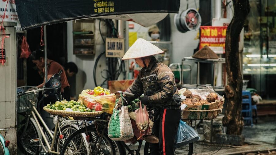 A street market vendor in Hanoi wearing a conical hat behind stacks of fruit and vegetables
