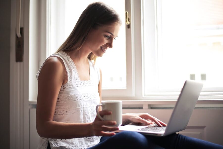 A woman with coffee using a laptop next to a window