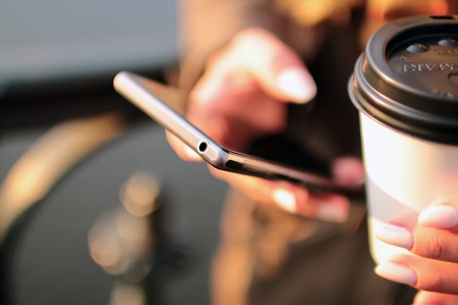 A person scrolling on a smartphone with a takeaway coffee in hand